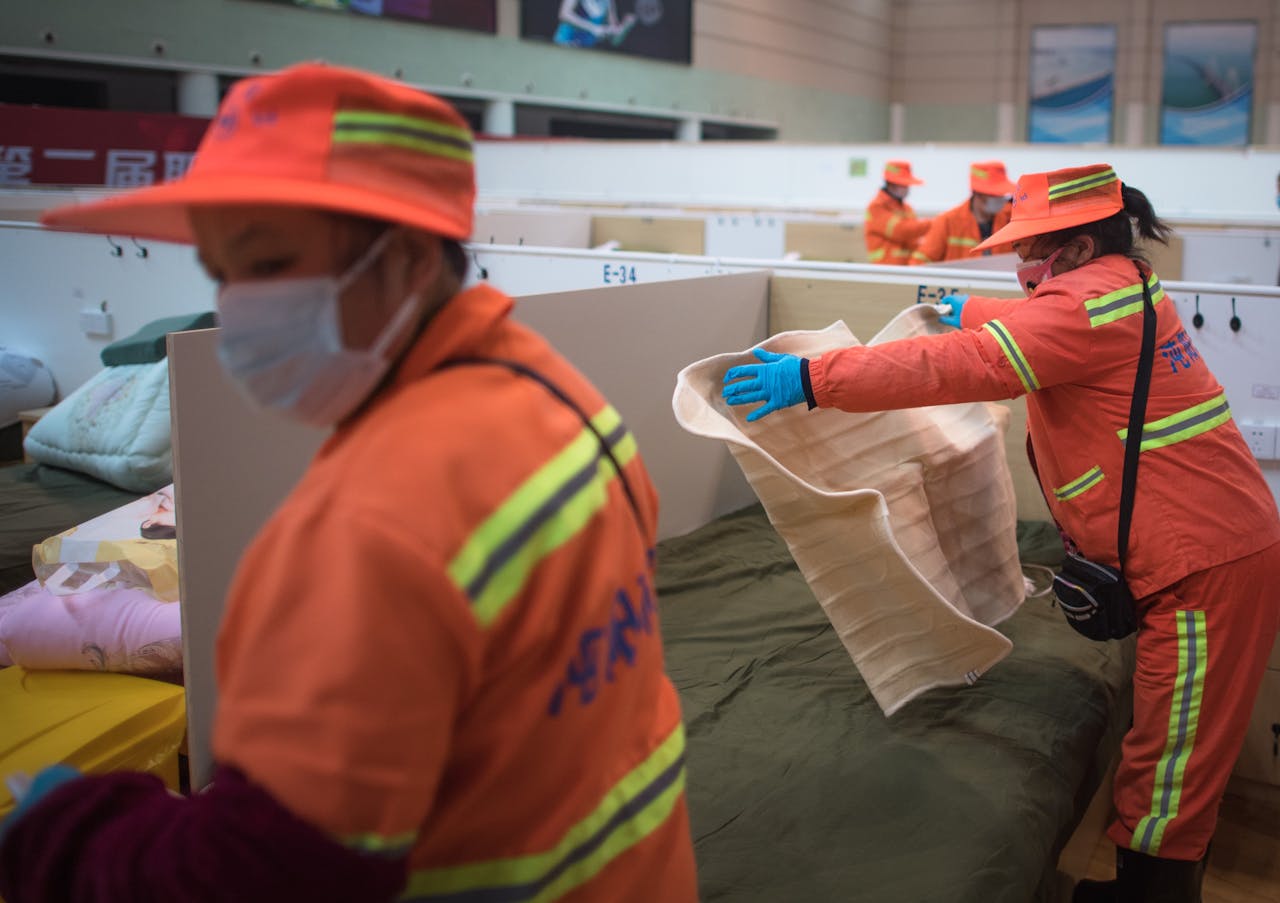 Emergency workers setting up temporary beds at an indoor facility wearing safety gear.